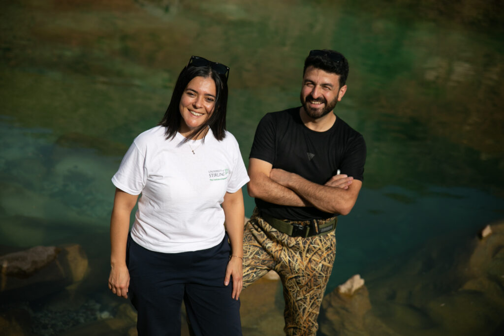 A photograph of a woman in a white t-shirt and black trousers, next to a man in a black t-shirt and green trousers. They are smiling in front of a river on a sunny day.