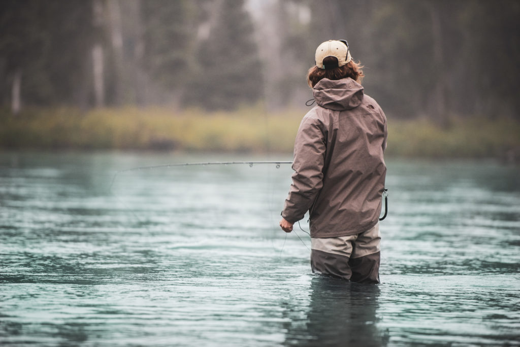 A photograph of a male angler in a beige hat and jacket standing in freshwater up to his knees.