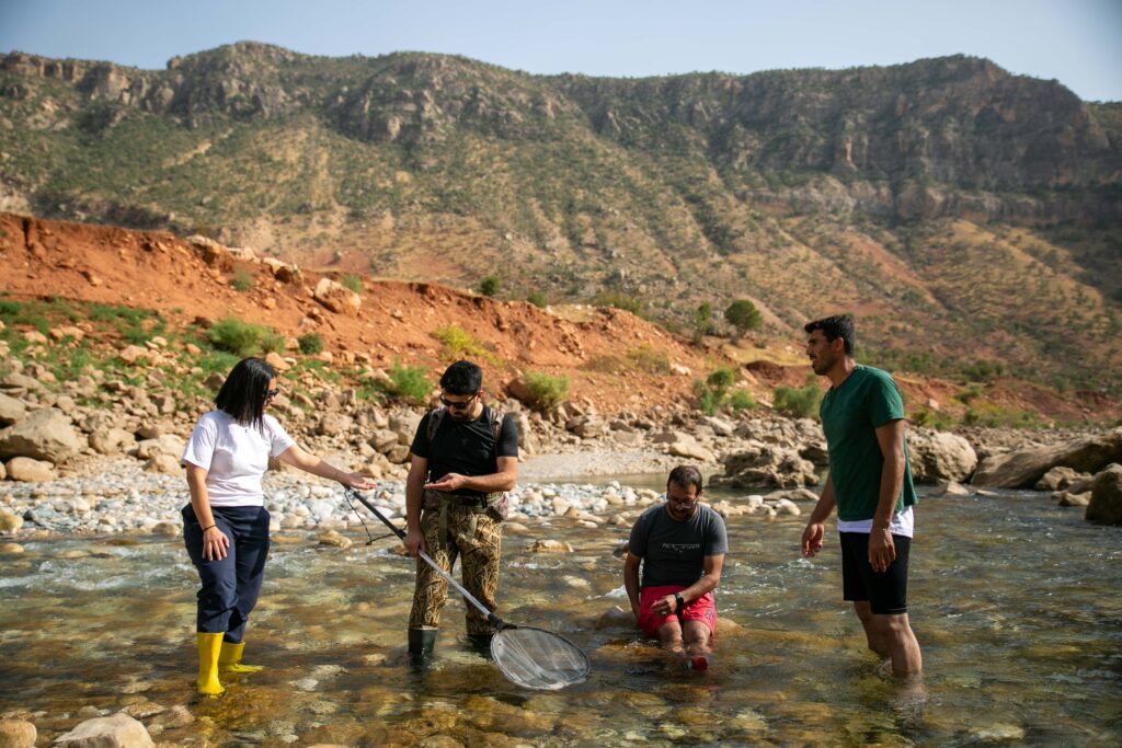 Photo of a woman and three men ankle deep in a river with mountains behind. One man is holding a big net.